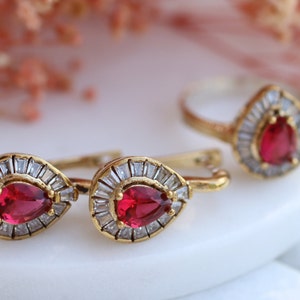 Może przedstawiać: Zestaw złotych kolczyków i pierścionka z czerwonymi kamieniami w kształcie łzy otoczonymi przez przezroczyste cyrkonie.