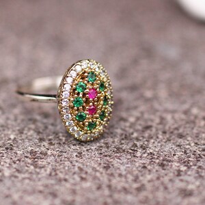 Pu&ograve; includere: Un anello ovale con una fascia color oro e un gruppo di pietre preziose. Il centro presenta pietre preziose rosa circondate da pietre preziose verdi e bianche. L'anello &egrave; posto su una superficie grigia strutturata.