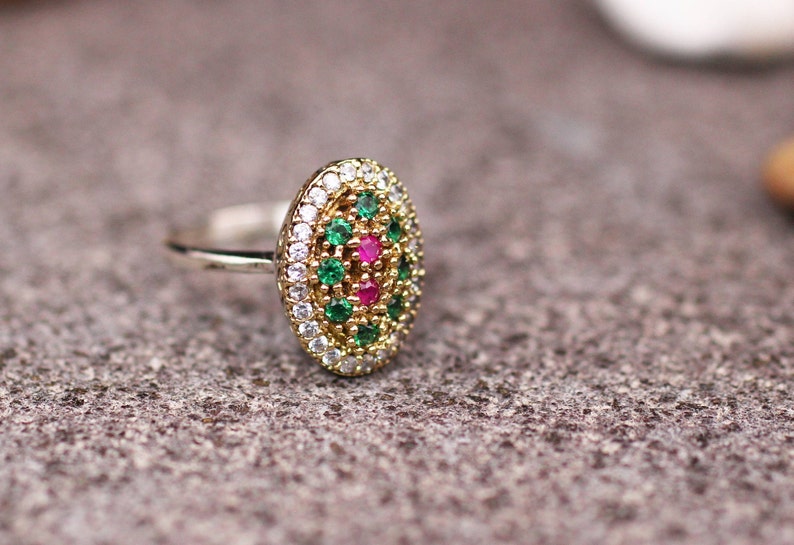 Pu&ograve; includere: Un anello ovale con una fascia color oro e un gruppo di pietre preziose. Il centro presenta pietre preziose rosa circondate da pietre preziose verdi e bianche. L'anello &egrave; posto su una superficie grigia strutturata.