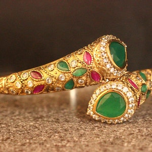 May include: A gold-toned bangle bracelet with a snake design. The bracelet is adorned with green and red gemstones and clear rhinestones.