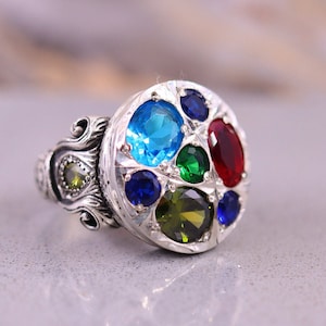 May include: A silver ring with a round, multi-coloured gemstone setting. The ring features a variety of blue, green, and red gemstones.
