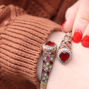 Może przedstawiać: Złota bransoletka typu bangle z czerwonymi i różowymi kamieniami szlachetnymi. Bransoletka jest otwarta i ma ozdobny wzór.