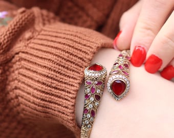 Pode incluir: Um bracelete de ouro com pedras preciosas vermelhas e rosadas. O bracelete é aberto e tem um design decorativo.