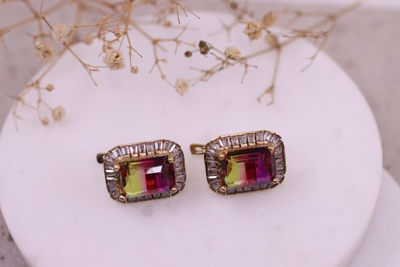 Puede incluir: Un par de pendientes dorados con piedras preciosas rectangulares multicolores. Cada pendiente presenta una piedra central con tonos rojos, amarillos y verdes, rodeada de peque&ntilde;os cristales transparentes. Los pendientes est&aacute;n sobre un fondo blanco.
