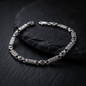 May include: A silver chain bracelet featuring alternating cylindrical links with an intricate woven pattern and smaller connector links, each set with a small, round, vibrant green gemstone. The bracelet includes a secure metal lobster clasp.