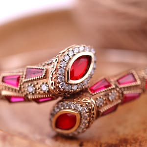May include: A gold snake-shaped bracelet featuring red and clear gemstones. The bracelet is intricately detailed with a delicate design.