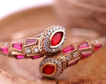 Puede incluir: Un brazalete de serpiente de oro con piedras preciosas rojas y transparentes. El brazalete está intrincadamente detallado con un diseño delicado.