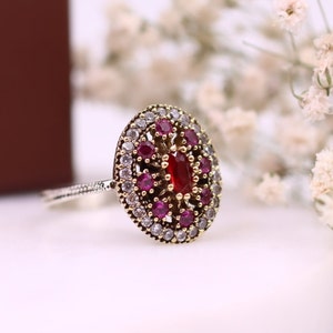 May include: A gold ring featuring a large oval red gemstone, encircled by smaller red and white gemstones.