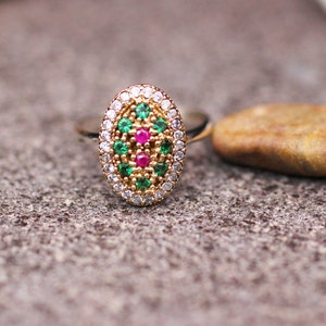 Pu&ograve; includere: Anello dorato con un elemento centrale ovale decorato con pietre verdi, rosse e bianche. Accanto all'anello, su una superficie grigia strutturata, c'&egrave; una piccola pietra liscia e marrone. Gioielli.