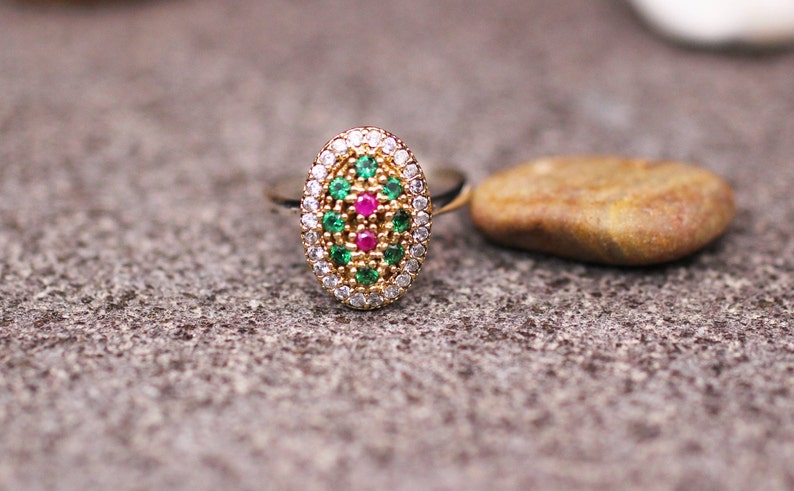 Pu&ograve; includere: Anello dorato con un elemento centrale ovale decorato con pietre verdi, rosse e bianche. Accanto all'anello, su una superficie grigia strutturata, c'&egrave; una piccola pietra liscia e marrone. Gioielli.