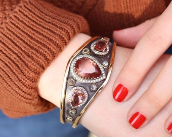 Puede incluir: Un brazalete de plata y oro con tres piedras preciosas en forma de lágrima. Las piedras preciosas son de color rosa claro y están rodeadas de piedras más pequeñas y transparentes. El brazalete está en la muñeca de una persona.