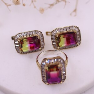 Puede incluir: Un conjunto de joyas con un anillo y pendientes a juego. Cada pieza tiene una piedra preciosa rectangular multicolor con un marco dorado y un halo de peque&ntilde;as piedras transparentes. El anillo y los pendientes se exhiben sobre una superficie blanca.