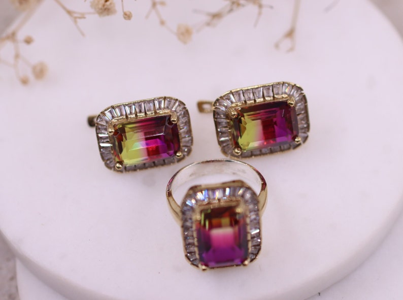 Puede incluir: Un conjunto de joyas con un anillo y pendientes a juego. Cada pieza tiene una piedra preciosa rectangular multicolor con un marco dorado y un halo de peque&ntilde;as piedras transparentes. El anillo y los pendientes se exhiben sobre una superficie blanca.