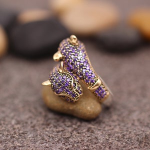K&ouml;nnte beinhalten: Ein goldfarbener Ring mit einem Panther-Design. Der Kopf und der K&ouml;rper des Panthers sind mit kleinen, lila Edelsteinen besetzt. Der Ring liegt auf einem kleinen, hellen Stein, mit einem verschwommenen Hintergrund aus anderen Steinen.