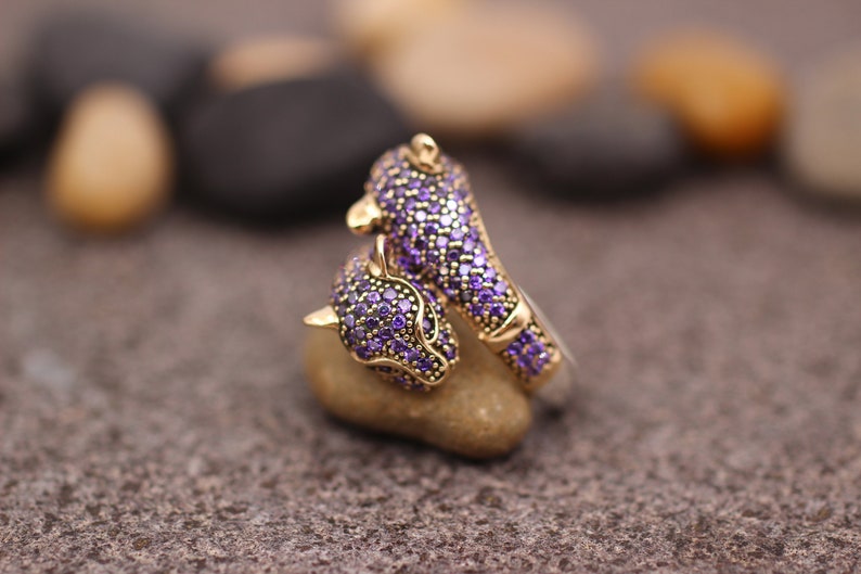 K&ouml;nnte beinhalten: Ein goldfarbener Ring mit einem Panther-Design. Der Kopf und der K&ouml;rper des Panthers sind mit kleinen, lila Edelsteinen besetzt. Der Ring liegt auf einem kleinen, hellen Stein, mit einem verschwommenen Hintergrund aus anderen Steinen.