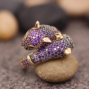 K&ouml;nnte beinhalten: Ein goldfarbener Ring mit einem stilisierten Panther-Design. Der Panther ist mit kleinen, runden, lila Edelsteinen besetzt. Der Ring liegt auf einem kleinen, abgerundeten Stein vor einem unscharfen Hintergrund.