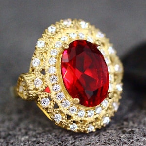 May include: A gold ring with a large, oval-shaped red gemstone surrounded by smaller, clear gemstones.
