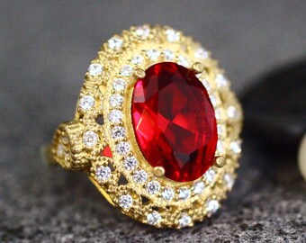 Könnte beinhalten: Ein goldener Ring mit einem großen, ovalen roten Edelstein, der von kleineren, klaren Edelsteinen umgeben ist.