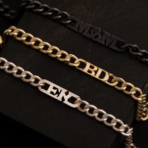 Könnte beinhalten: Drei Kettenarmbänder in Gold, Silber und Schwarz mit personalisierten Namensschildern. Das goldene Armband trägt den Namen "BDD", das silberne Armband den Namen "EX" und das schwarze Armband den Namen "M&M".