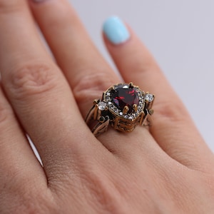 Op de afbeelding: Een gouden ring met een grote, hartvormige, rode edelsteen omringd door kleinere, heldere edelstenen.