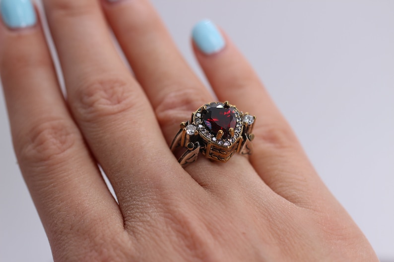 Op de afbeelding: Een gouden ring met een grote, hartvormige, rode edelsteen omringd door kleinere, heldere edelstenen.