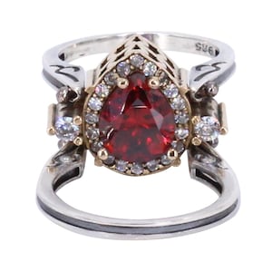 Op de afbeelding: Een zilveren en gouden ring met een grote peervormige rode edelsteen omringd door kleinere heldere edelstenen. De ring heeft een delicaat ontwerp met ingewikkelde details.