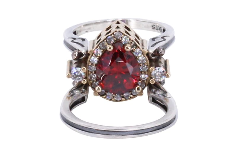 Op de afbeelding: Een zilveren en gouden ring met een grote peervormige rode edelsteen omringd door kleinere heldere edelstenen. De ring heeft een delicaat ontwerp met ingewikkelde details.