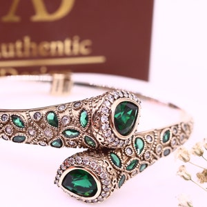 Pode incluir: Um bracelete de ouro com designs florais intrincados e duas grandes pedras preciosas verdes em forma de pêra. O bracelete é adornado com pedras preciosas menores e transparentes.