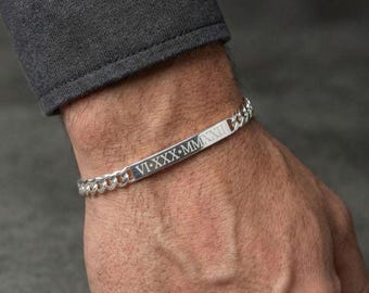 May include: A silver curb chain bracelet with a rectangular bar engraved with Roman numerals. The bracelet is on a wrist, and the chain links are visible. The bar has the Roman numerals VI-XXX-MMXII.