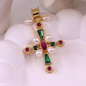 以下が含まれることがあります： 中央に楕円形のルビー、エメラルド、真珠、小さなルビーをあしらった、金色のクロス型ペンダント。クロスは詳細なデザインで、白い背景にセットされています。