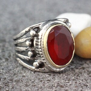 Op de afbeelding: Een zilveren ring met een rode edelsteen gezet in een gouden bezel. De ring heeft een gedetailleerd ontwerp met meerdere banden en kleine kralen.