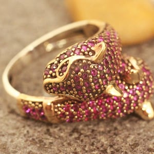 以下が含まれることがあります： ピンクの宝石のデザインが施された、動物が丸まっているような形の金の指輪。指輪には小さな輝くピンクの石が埋め込まれています。