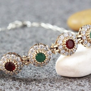 Puede incluir: Un brazalete de plata con una cadena de oro y cinco abalorios redondos. Cada abalorio está decorado con pequeñas piedras blancas y una piedra central más grande. Las piedras centrales son rojas, verdes y rojas.