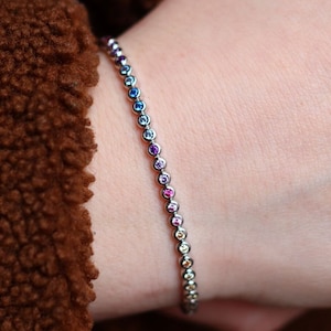 May include: A delicate bracelet featuring a series of small, colorful gemstones. The gemstones are arranged in a gradient of colors, transitioning from blue to purple, pink, and yellow. The bracelet is made of a silver-colored metal and is worn on a wrist.