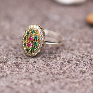 Pu&ograve; includere: Anello dorato con un elemento centrale ovale ornato da piccole pietre preziose rotonde verdi, bianche e rosse. L'anello &egrave; posizionato su una superficie grigia strutturata.