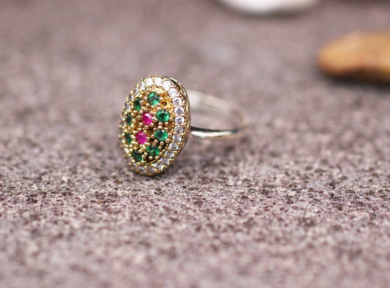Pu&ograve; includere: Anello dorato con un elemento centrale ovale ornato da piccole pietre preziose rotonde verdi, bianche e rosse. L'anello &egrave; posizionato su una superficie grigia strutturata.