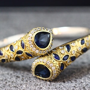 May include: A gold-toned cuff bracelet with intricate detailing. The bracelet features large, dark blue teardrop-shaped gemstones surrounded by smaller white gemstones. Additional blue gemstones are set along the band.