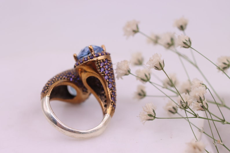 May include: A silver and gold ring with a large blue gemstone surrounded by smaller purple gemstones.