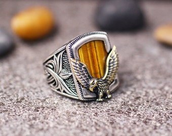 May include: A silver ring with an eagle design and a tiger's eye stone. The eagle is perched on a shield-shaped setting with intricate details.
