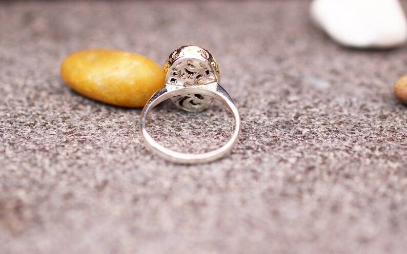 Pu&ograve; includere: Anello d'argento con castone ovale dettagliato con filigrana. L'anello &egrave; appoggiato su una superficie grigia strutturata, con una pietra gialla e una pietra bianca sullo sfondo. La fascia dell'anello &egrave; liscia e lucida.