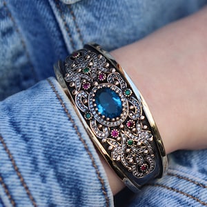 以下が含まれることがあります： 大きな青い宝石が、より小さな赤と緑の宝石で囲まれた、銀と金のバングルブレスレット。ブレスレットは、複雑な花の模様をしています。