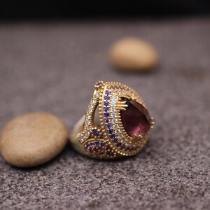 Puede incluir: Un anillo de oro con una gran piedra preciosa p&uacute;rpura rodeada de piedras m&aacute;s peque&ntilde;as transparentes y p&uacute;rpuras.