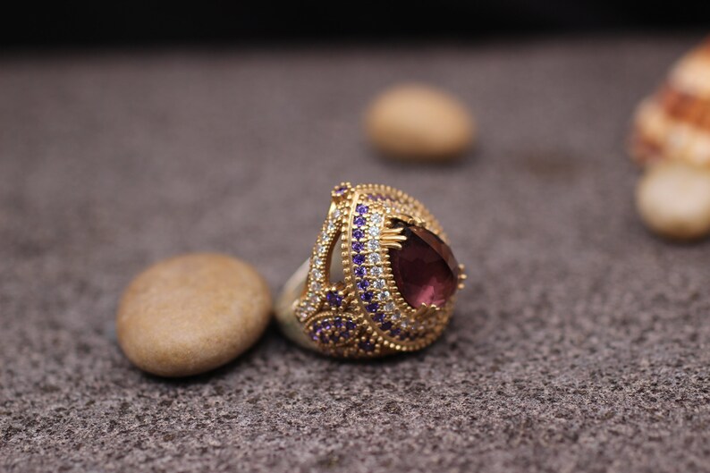 Puede incluir: Un anillo de oro con una gran piedra preciosa p&uacute;rpura rodeada de piedras m&aacute;s peque&ntilde;as transparentes y p&uacute;rpuras.