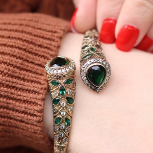 May include: A gold bangle bracelet with green and clear stones. The bracelet is open and has a curved design.