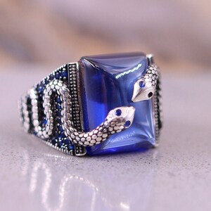 May include: A silver ring featuring a large blue gemstone and two snake designs on the side. The snakes are facing each other and have blue eyes.