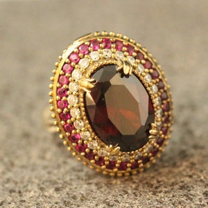 Op de afbeelding: Een gouden ring met een grote, ovale, donkerrode edelsteen omringd door een halo van kleinere, witte en rode edelstenen.