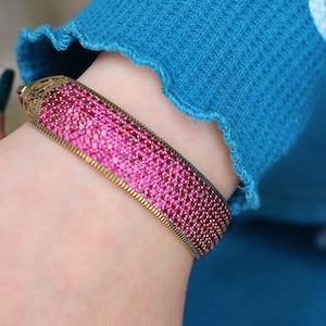 May include: A gold bangle bracelet with a pink, sparkly design. The bracelet is made of small, round stones that are set close together.