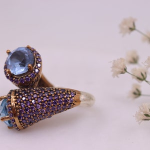 May include: A silver ring with two blue gemstones set in a gold-tone setting. The ring is adorned with small purple gemstones.