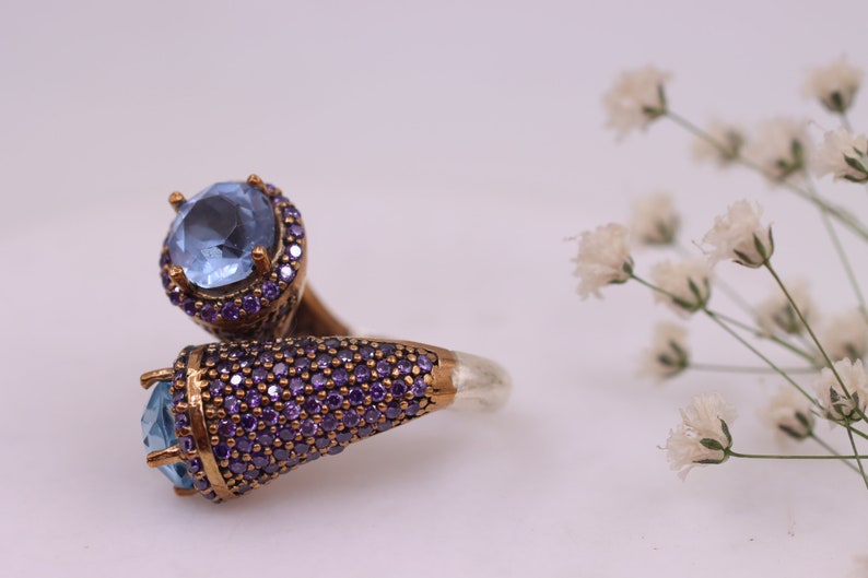 May include: A silver ring with two blue gemstones set in a gold-tone setting. The ring is adorned with small purple gemstones.