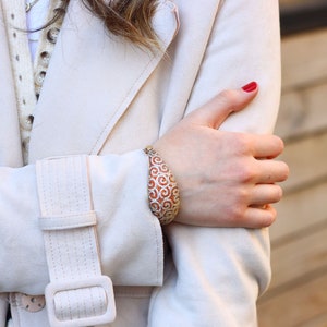 K&ouml;nnte beinhalten: Nahaufnahme eines Frauenhandgelenks, das ein gold- und orangefarbenes Armband mit einem Spiralmuster tr&auml;gt. Das Armband ist aus Metall und hat ein gro&szlig;es, ovales Design. Die Frau tr&auml;gt einen wei&szlig;en Mantel und hat roten Nagellack.